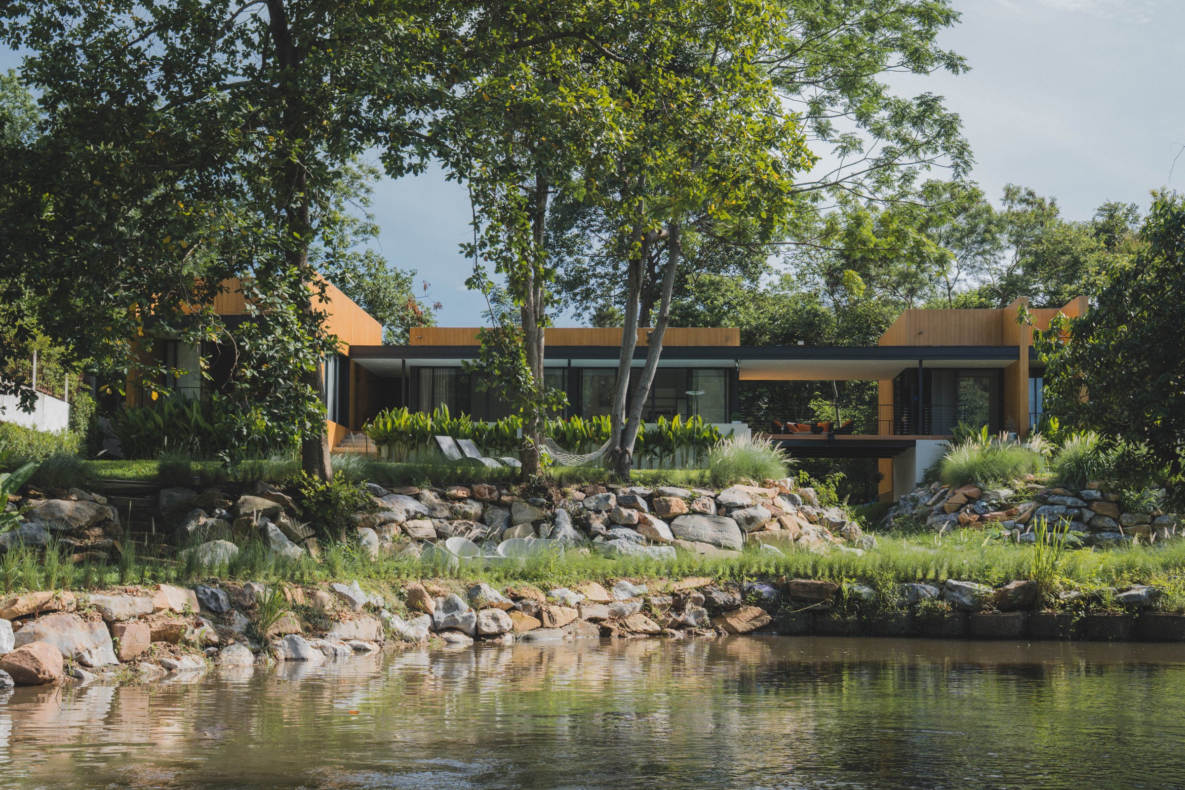 Timber-clad villa seen across a stream and boulder bank, under tall trees