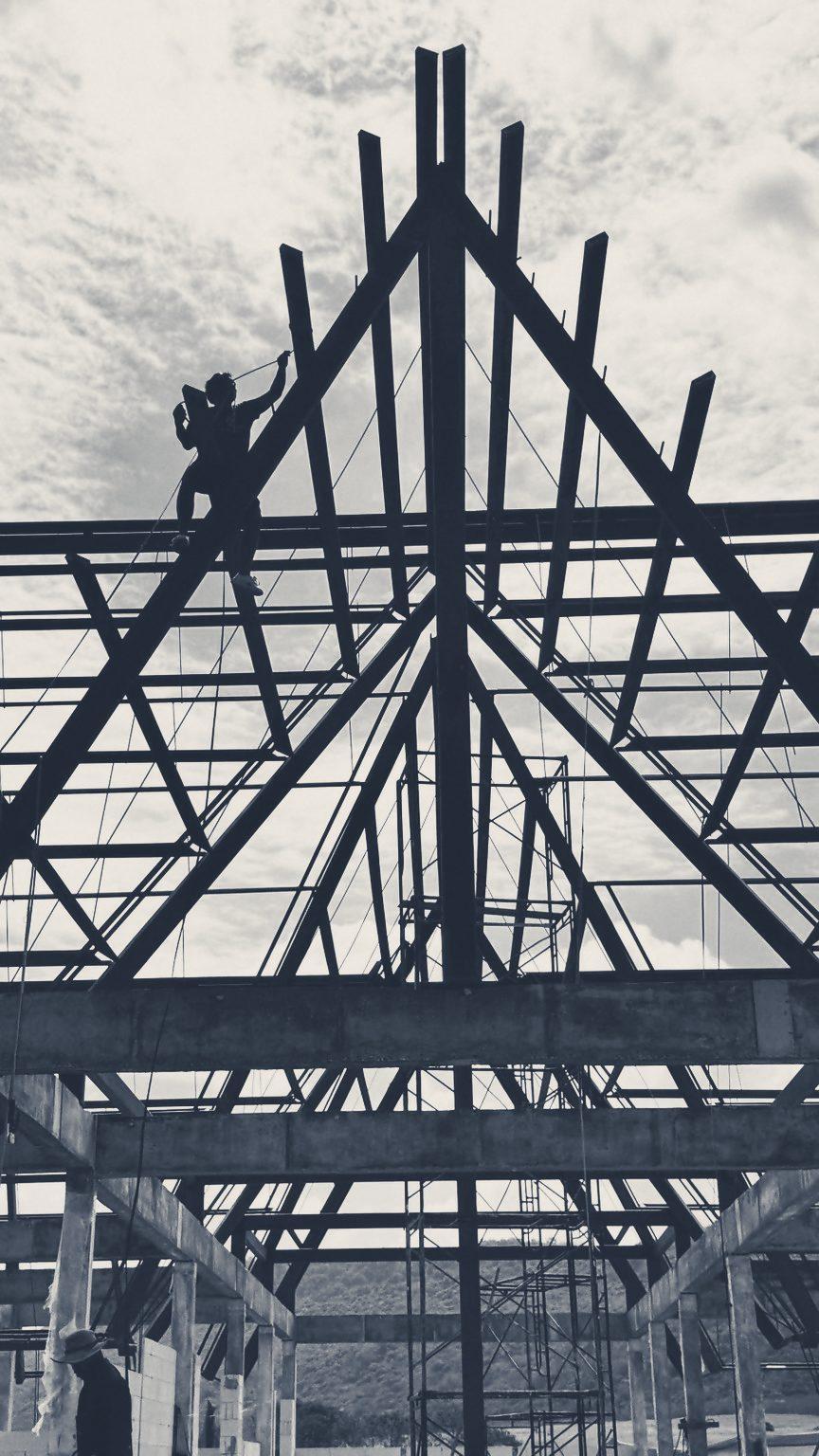 The steel roof frame of the Club House against a cloudy sky, with workers up on the frame