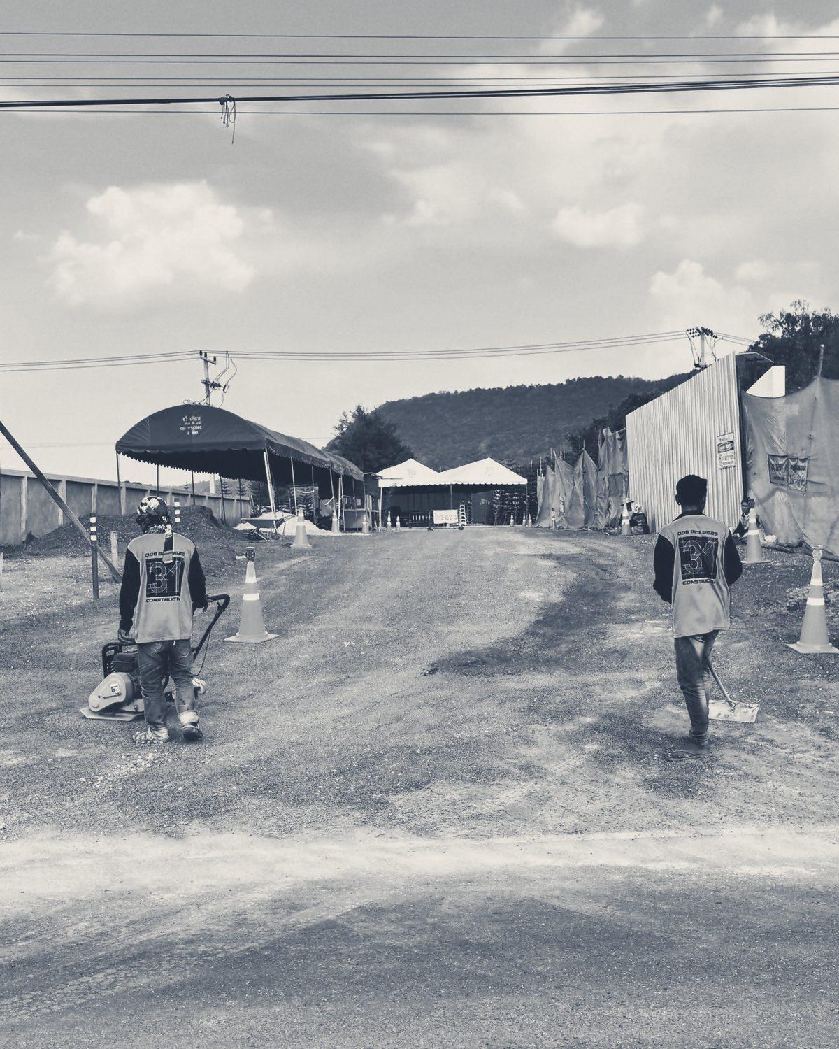 Two CON31 crew in 31 vests walk onto the Main Gate site with their backs to the camera, one pushing a plate compactor, one carrying a shovel.