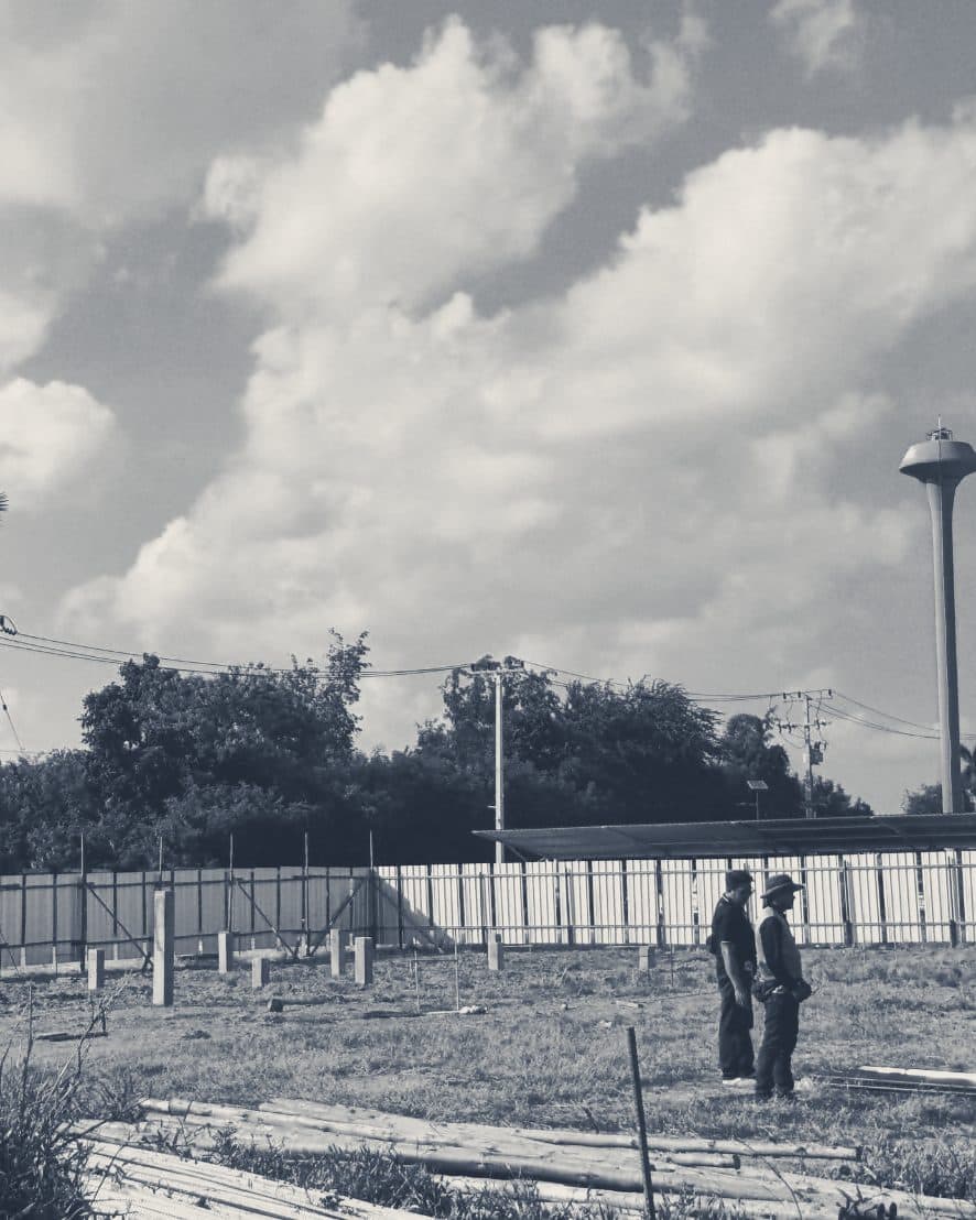 Two people standing on a cleared site before building starts, with a fence and trees behind