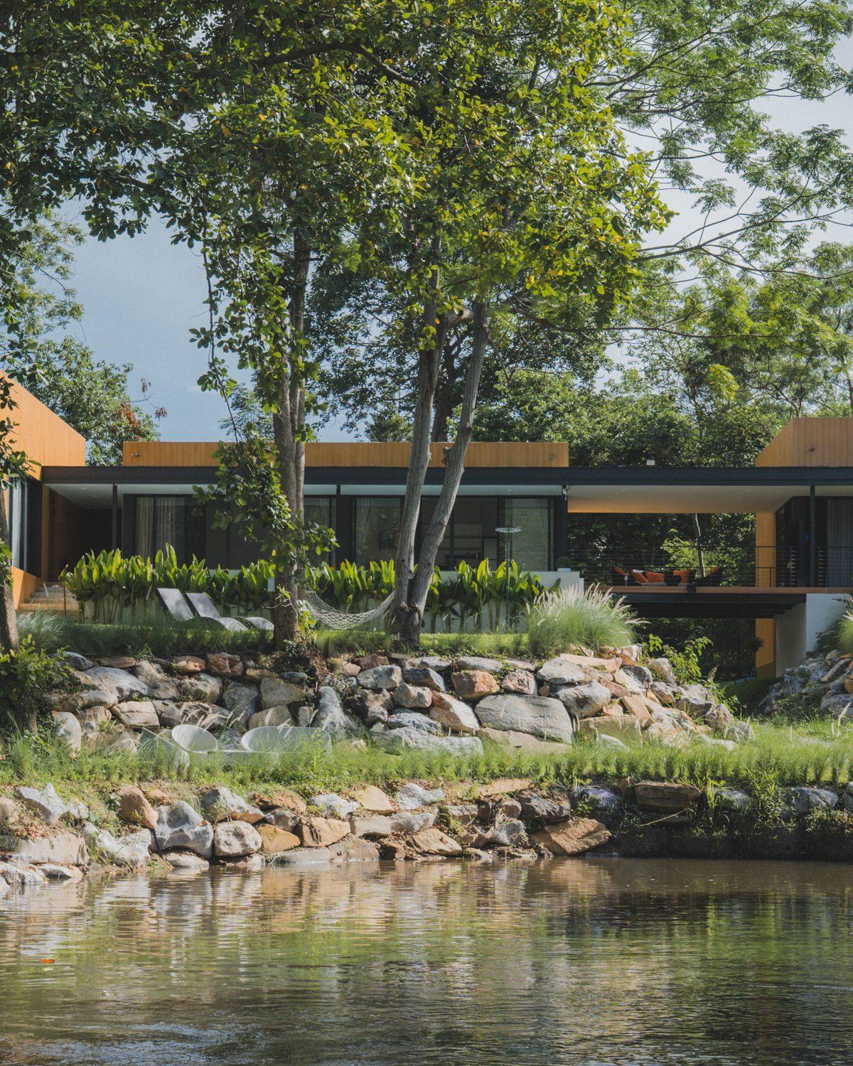 The timber-clad villa seen across a stream and a boulder bank, under tall trees