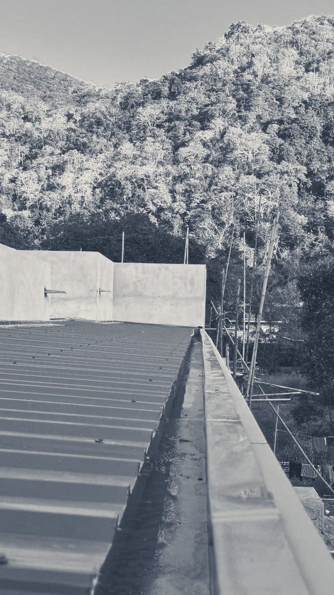 Metal roof sheets running down to the gutter beside a capped parapet, with bamboo scaffold and a wooded hill behind