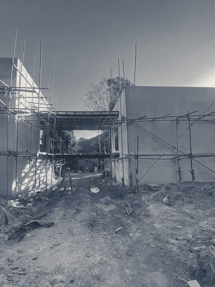 The villa’s two wings in bamboo scaffold and the steel frame of the bridge between them, seen from the dirt site track